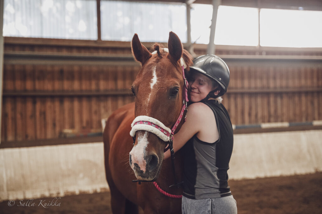 Kuvassa yksi henkilö halaa punaruskeaa hevosta, jolla on punainen, vaalean karvapehmusteen omaava riimu. Kuva on otettu Mennalan Ratsutilan maneesissa ja sen taustalla näkyy maneesin seinä ja puomeja